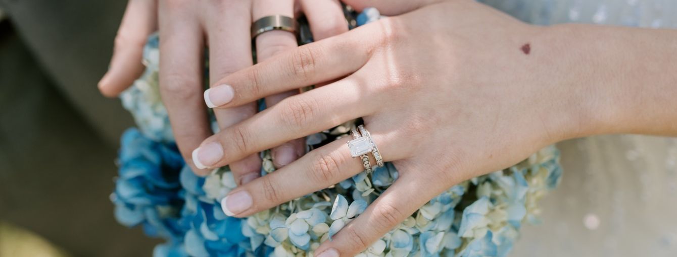 A couple's hands are together over the top of her bouquet. 